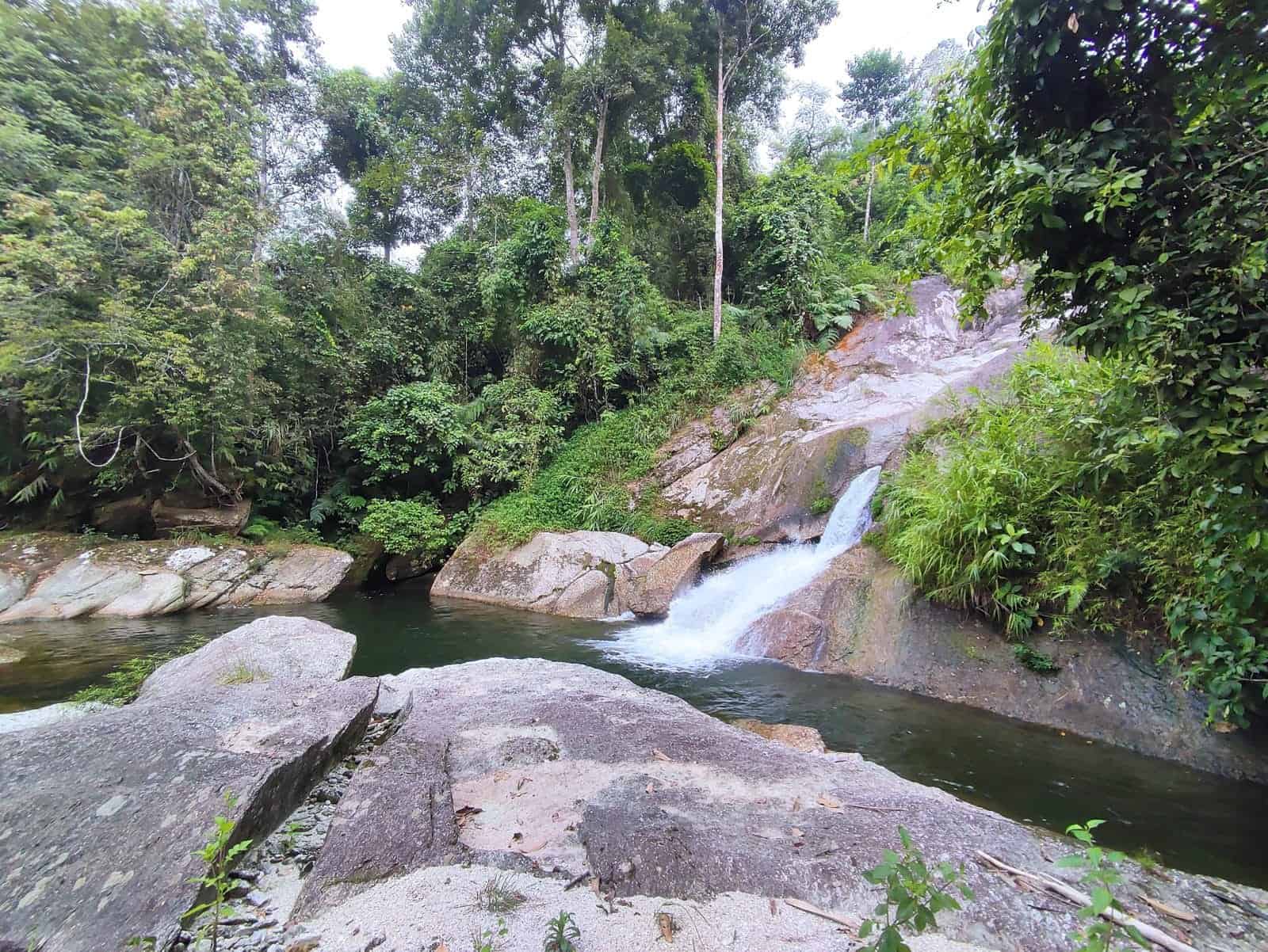 Lung Vai Waterfall spilling into a green pool beneath tall forest on the 1-day Khuoi My trek, Ha Giang