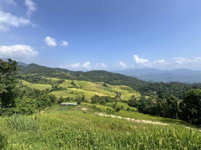 Terraced rice fields at Na Thac ripening above the welcome gate, with the forested ridges of Tay Con Linh and the karst peaks of Ha Giang beyond