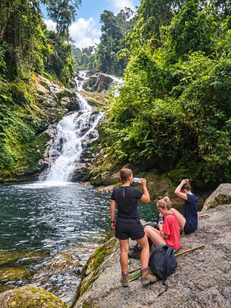 Trekkers resting on a rock by a waterfall pool